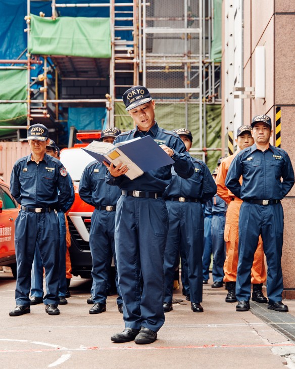 Tokyo Fire Department firefighters in blue uniforms and white caps stand at attention during morning shift change at Ikebukuro Fire Station.
