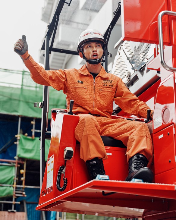 Ladder operator Yuki Wada in orange protective gear sitting in elevated equipment, giving a thumbs-up to his crew below.
