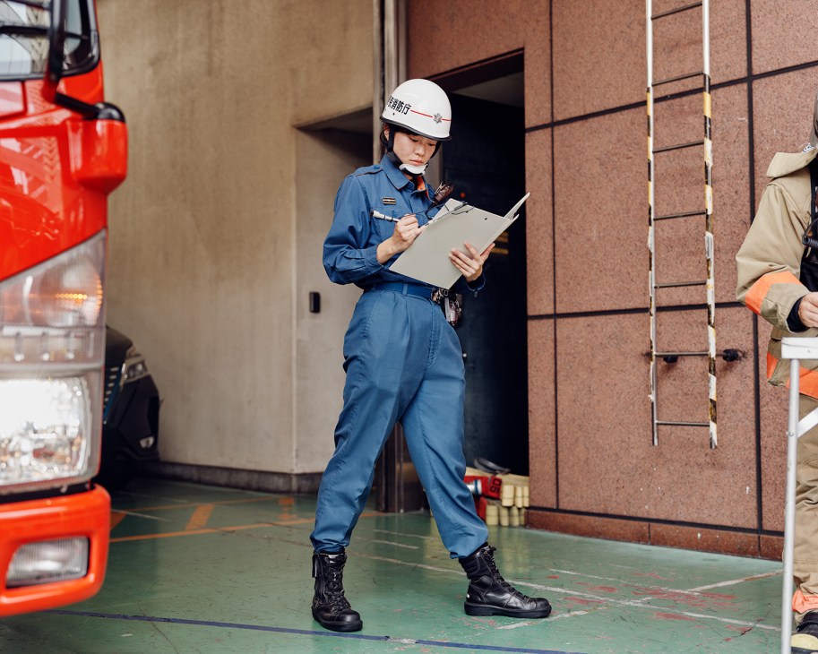 Female firefighter in blue uniform and white helmet holding clipboard during drill outside brick building