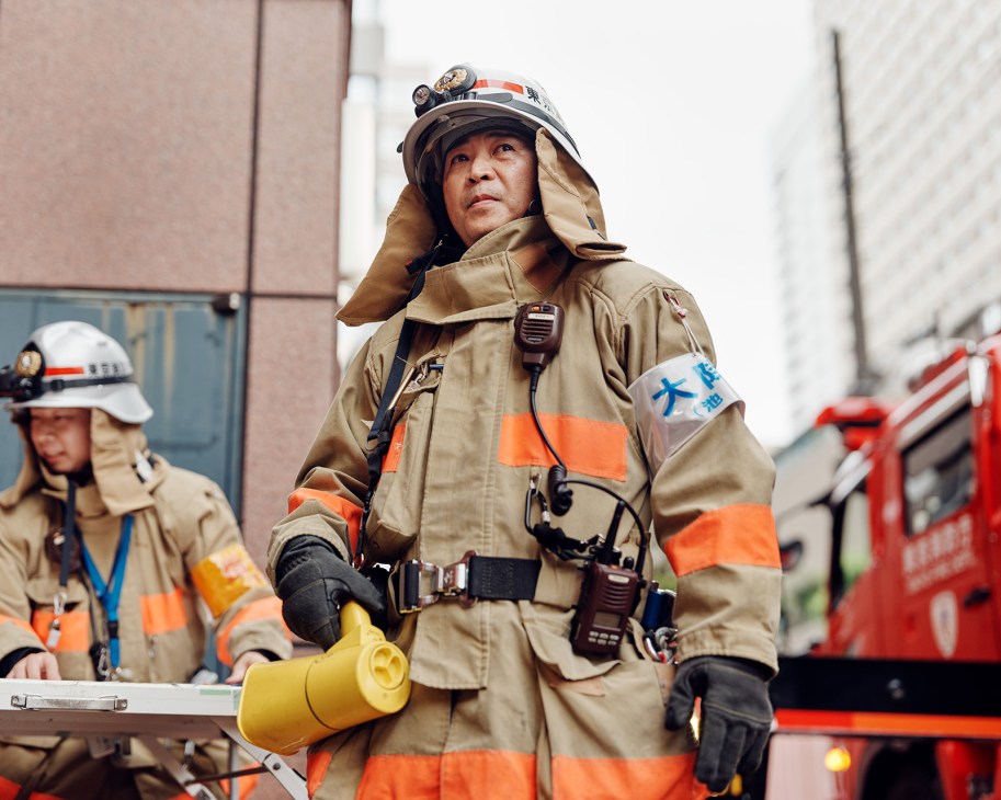 Tokyo Fire Department firefighter in tan protective gear coordinating a ladder-rescue drill with a megaphone.