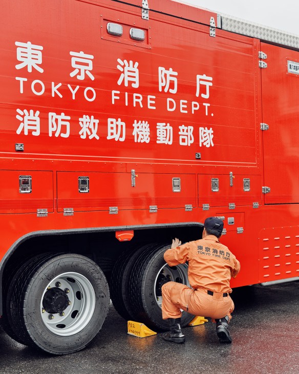 Tokyo Fire Department firefighter in orange hazmat suit crouching beside red fire truck wheel.