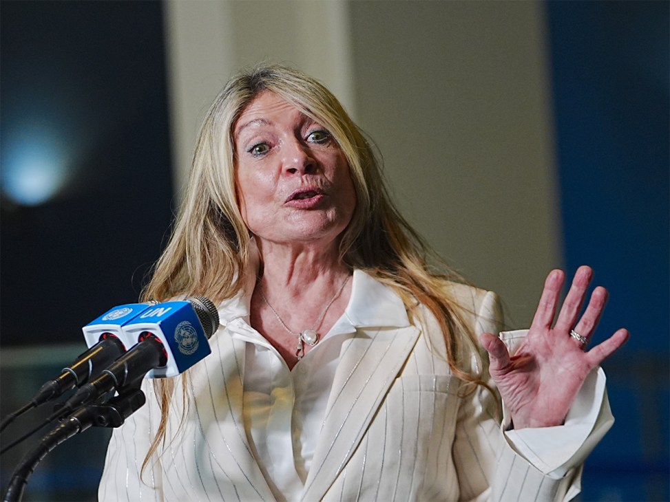 Woman in cream pinstriped blazer speaking at microphones during a public address.