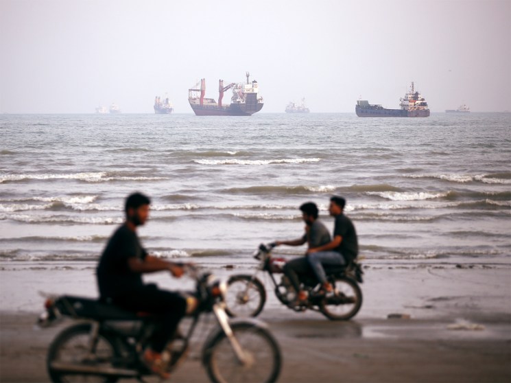 Merchant ships anchored in the Strait of Hormuz with people on motorcycles on the beach in foreground.