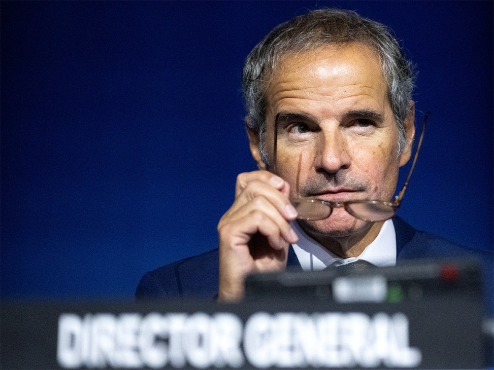 Man in dark suit and white shirt wearing a headset microphone against a blue background.