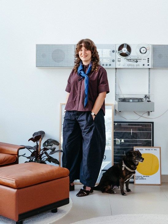 Woman standing in office with vintage audio equipment and reel-to-reel recorder behind her, alongside orange leather furniture and black dog.