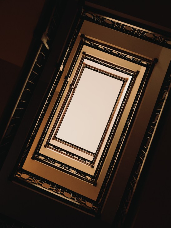 Looking up through a rectangular stairwell with ornate black and tan metal railings receding toward a bright opening.