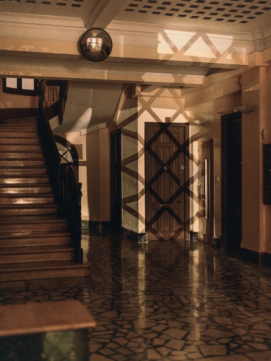 Sunlit entrance hall with geometric shadows cast through latticed railings onto polished floor