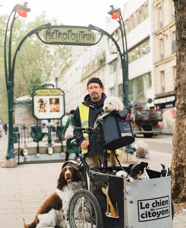 Alain Lambert rides a cargo bike with three dogs along a Paris street near a Metro entrance sign.