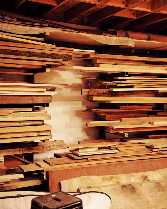 Stacked wooden planks of varying colors in a workshop, with exposed wooden ceiling beams above.