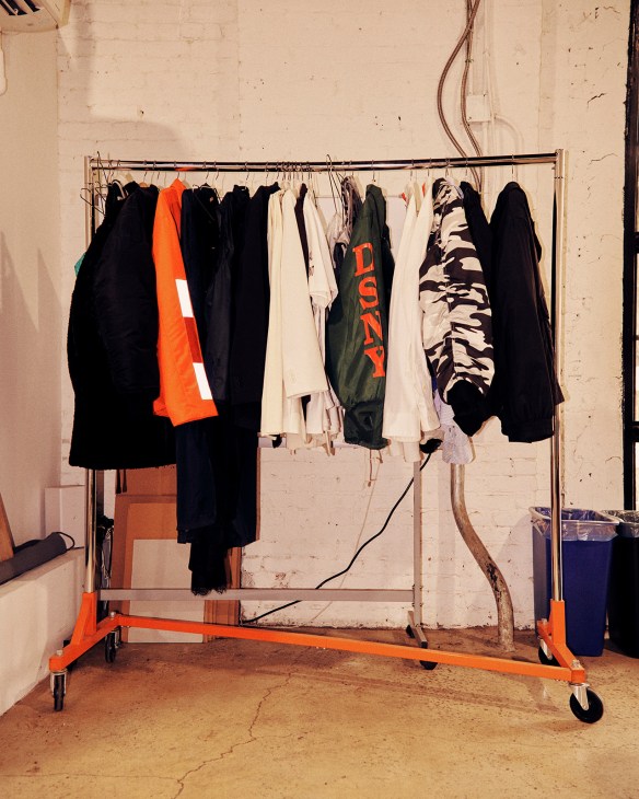 Clothing rack with black, white, and orange garments including a green jacket with orange text at Heron Preston's studio.