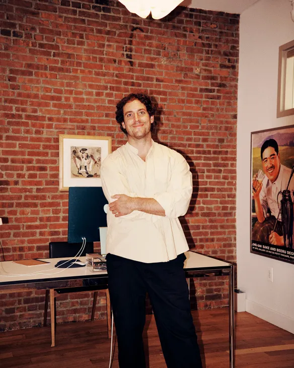 Man in cream shirt standing at desk in brick-walled apartment with exposed brick and artwork.