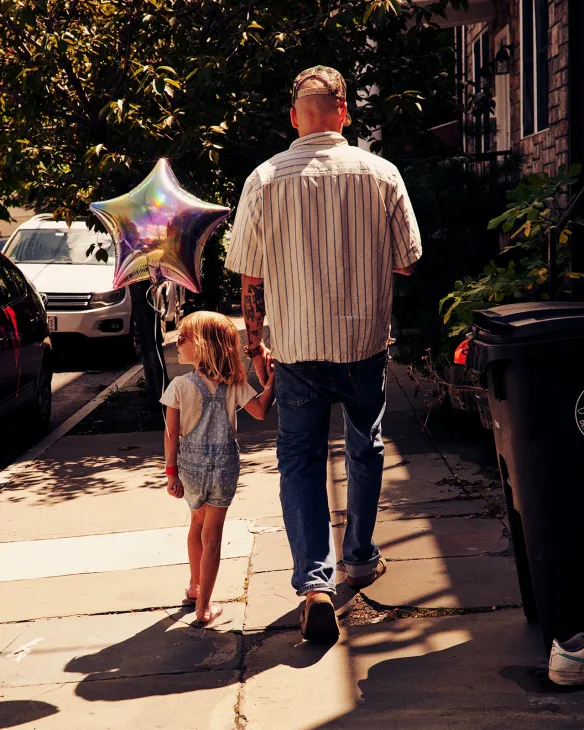 Adult and child walking on a sunny Red Hook street holding a colorful star balloon.