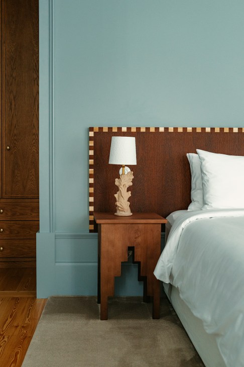 Bedroom with blue-green wall, brown wooden headboard with checkerboard border, wooden nightstand, and cream table lamp with leaf-shaped base.