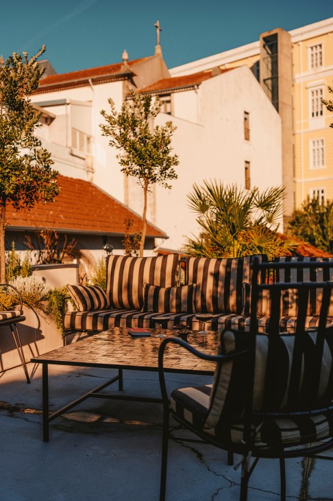 Striped outdoor lounge chairs and a metal table on a sunny rooftop terrace with cream-colored buildings and potted plants in the background.