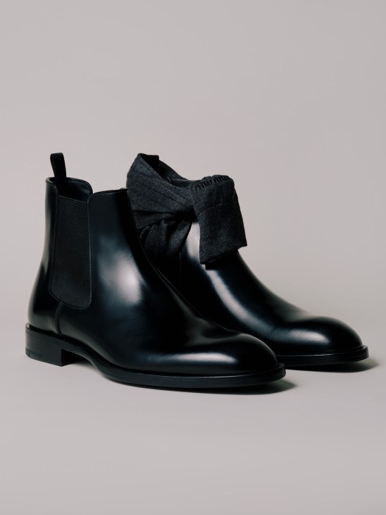Pair of black leather Chelsea boots with a fabric bow detail, styled with dark socks against a gray background.