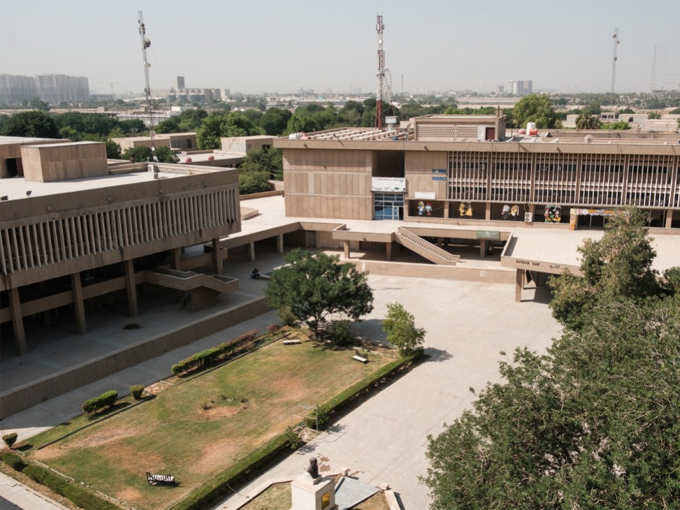 Brutalist concrete courtyard with modernist buildings, trees, and Baghdad cityscape in background.