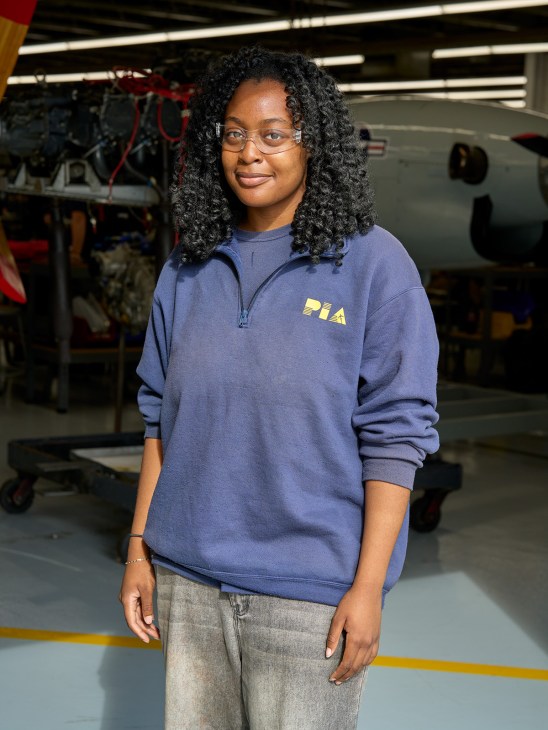 A PIA student in a blue sweatshirt stands in an aircraft hangar with maintenance equipment visible in the background.