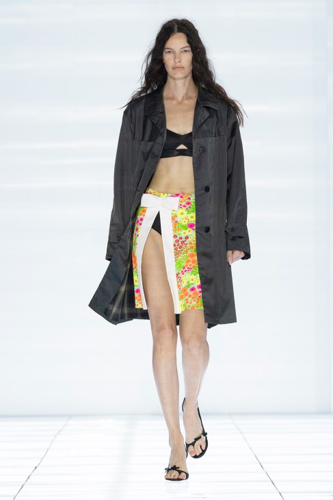 Model on runway wearing Prada spring/summer 2027 collection with colorful floral skirt, black crop top, and charcoal trench coat.