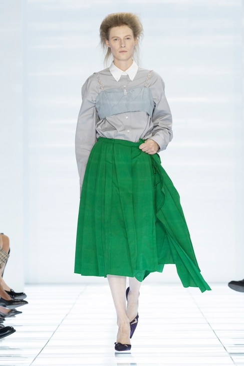 Model wearing gray shirt with layered blue overlay and full green pleated skirt on runway.