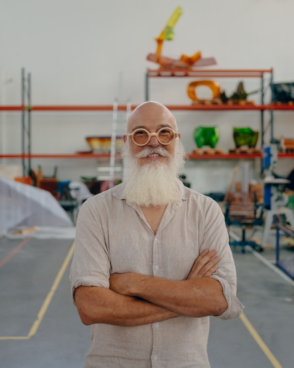 Guillaume Crédoz stands with arms crossed in his workshop, wearing glasses and a light shirt, with industrial shelving and colorful equipment visible behind him.