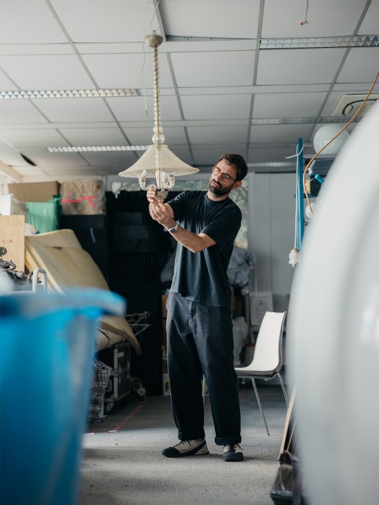 Man in glasses and black shirt holding a cream-colored pendant light fixture in a workshop warehouse.