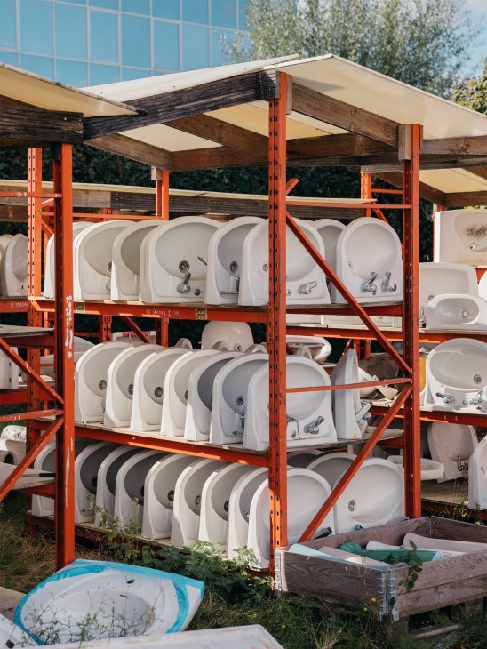 White bathroom sinks displayed on red metal industrial shelving in a warehouse courtyard.