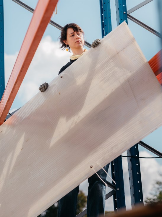 Woman holding a large light-colored wooden beam against a steel industrial frame structure.