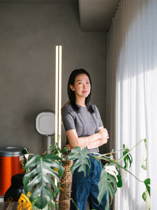 Woman standing indoors with arms crossed, surrounded by potted plants and a vertical light fixture