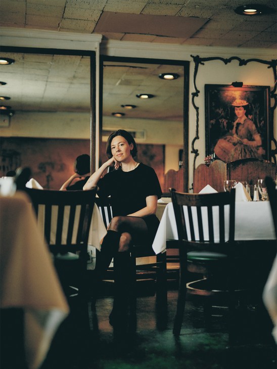 Woman in black shirt seated at a table in Gene's restaurant, Manhattan, with white tablecloth and portrait painting visible behind her.