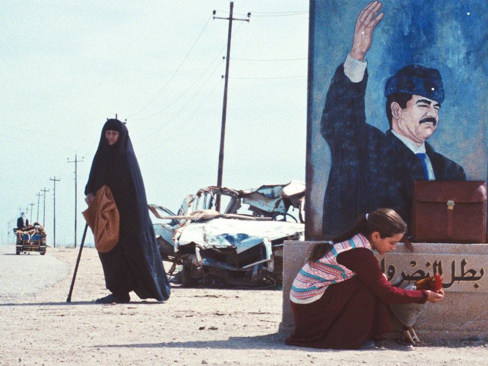 A woman and child stand before a mural of a man with raised fist in a desert landscape with utility poles.
