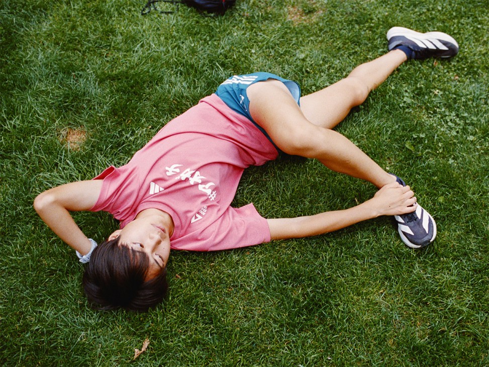Person lying on grass wearing pink shirt and blue shorts with athletic shoes.