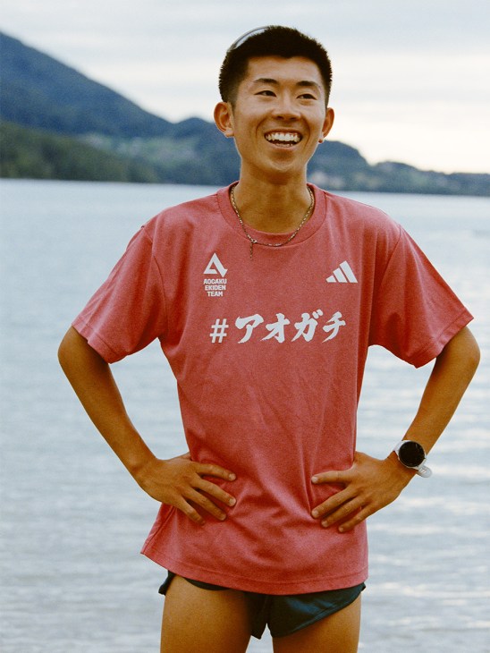 Young male runner wearing red Adidas Aoyama Gakuin University team shirt smiling by a lake with mountains in background