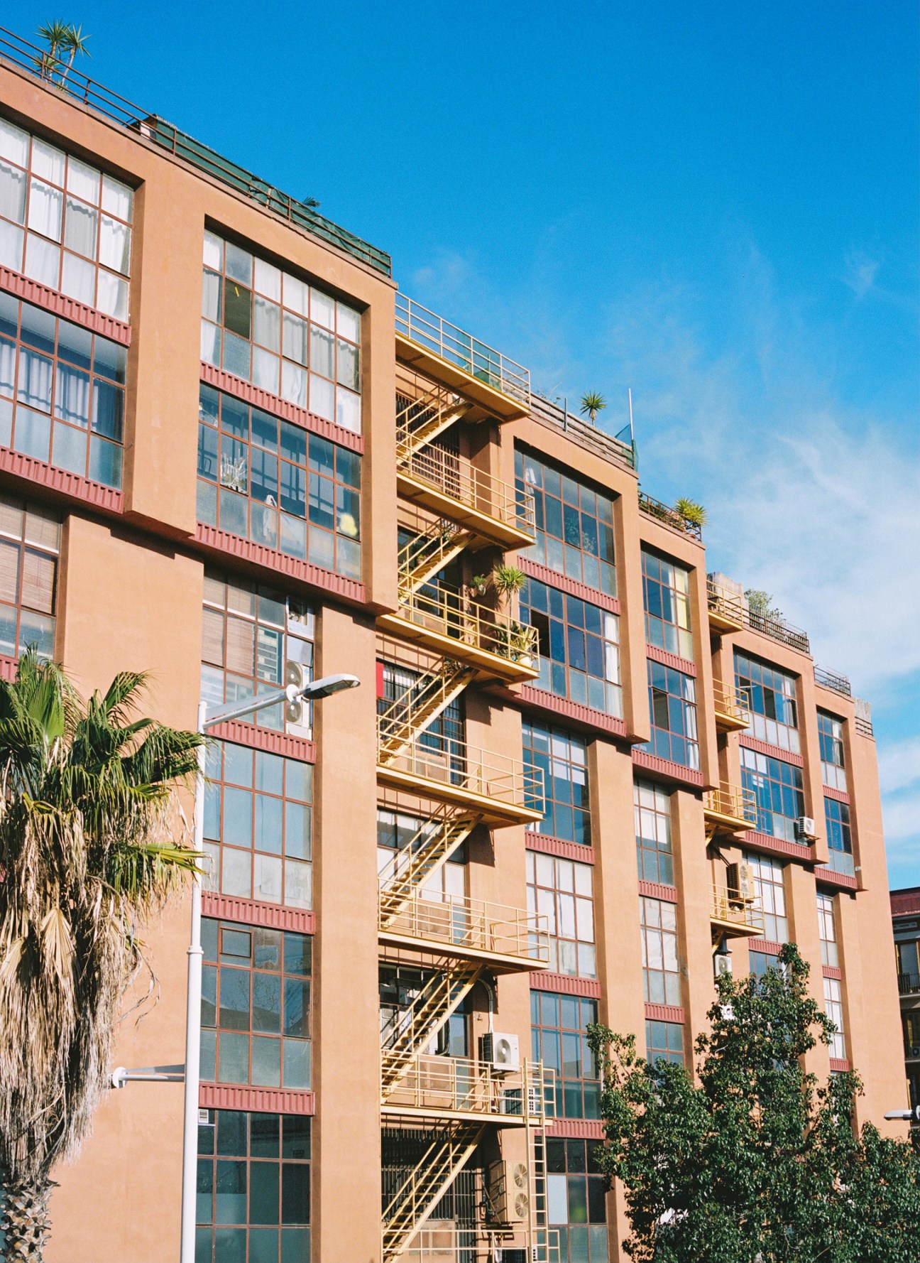 Terracotta-coloured building facade with large grid windows, red metal trim, and yellow external staircases against blue sky.