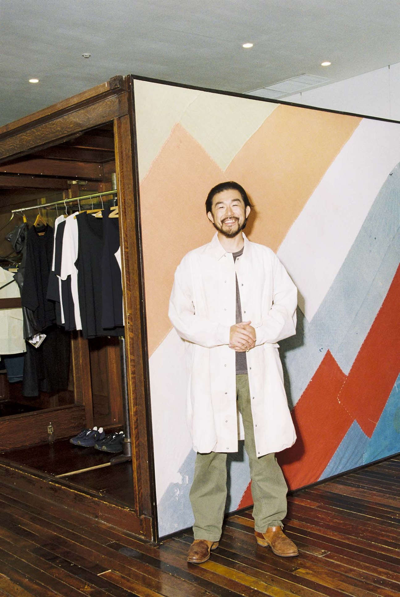 Takashi Takagi wearing a white shirt jacket over green trousers and brown shoes inside a Visvim store.