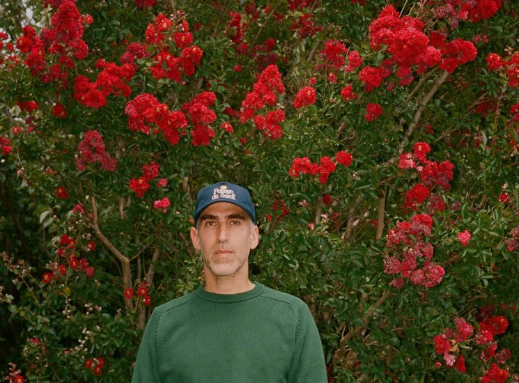 Brendon Babenzien wearing a green sweater and baseball cap standing in front of red flowering trees