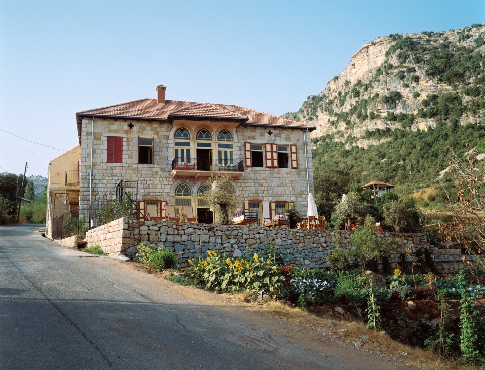 Traditional Lebanese stone house with red tile roof set against mountainous landscape with vegetation