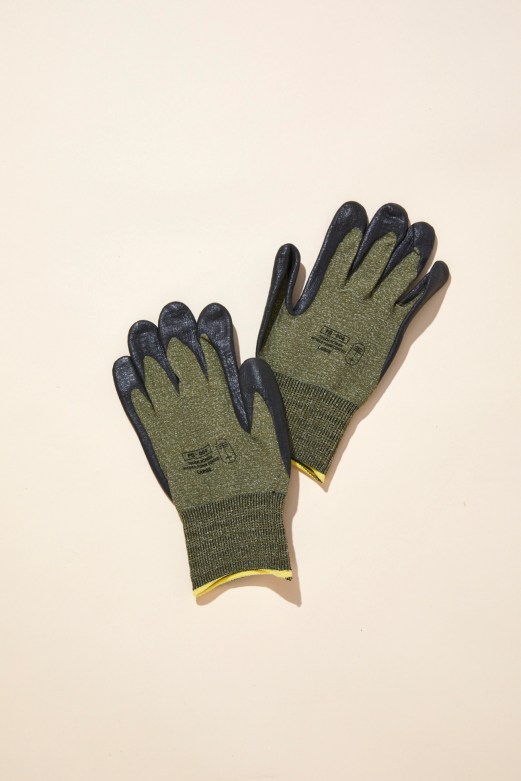 A pair of olive green work gloves with dark fingertips and yellow cuff trim laid on a beige surface