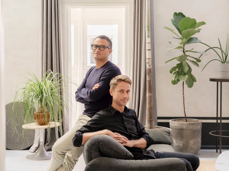 Architects Chris Briffa and Leroy Merks pose in a bright, minimalist interior space with plants and natural light.