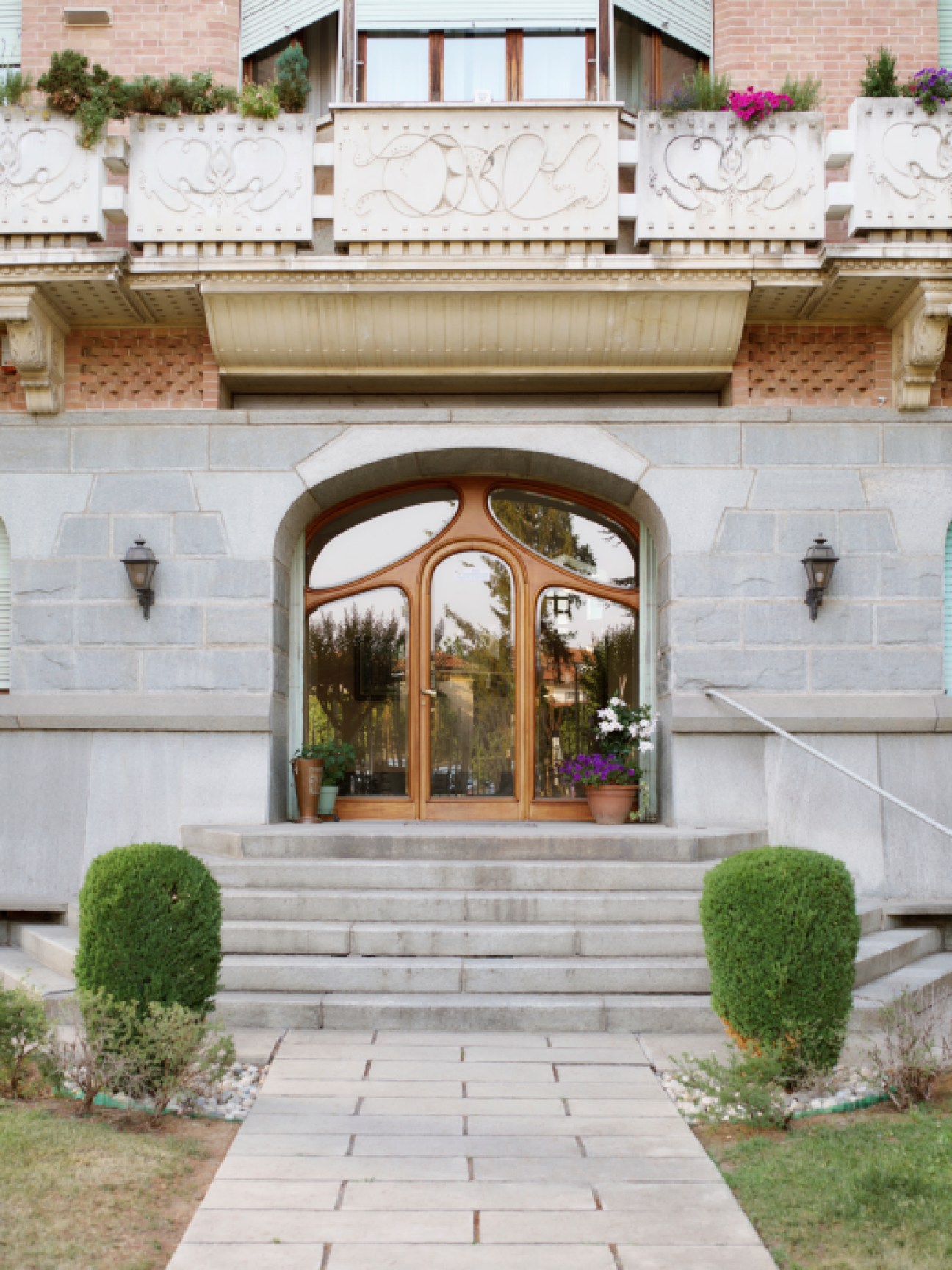 Ornate arched entrance with curved glass doors, decorative stonework, and manicured hedges flanking stone steps
