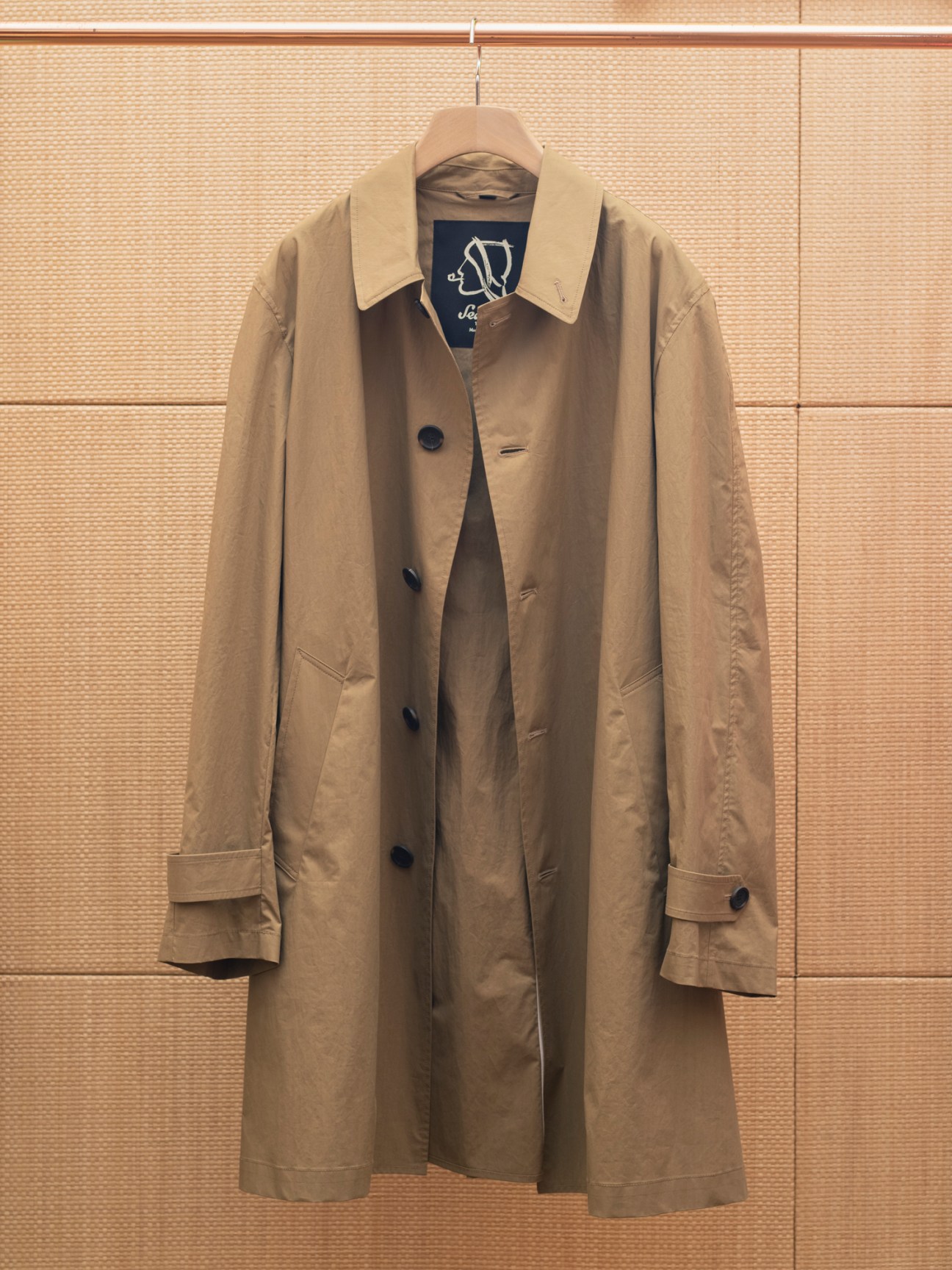 Tan Sealup Bretello trench coat hanging on wooden hanger against bamboo wall