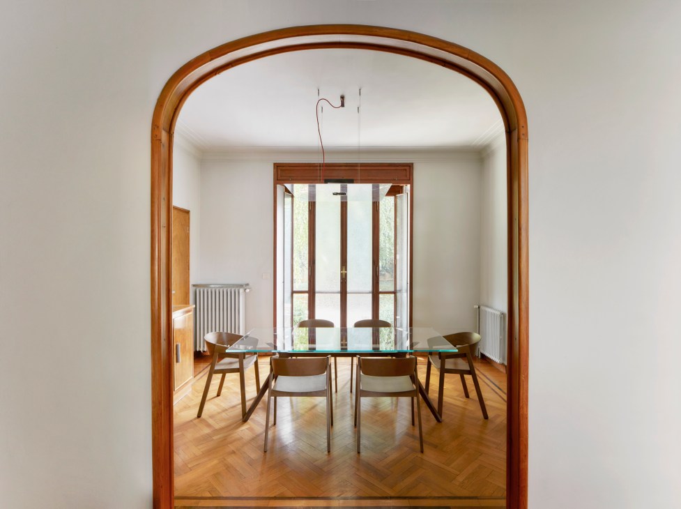A dining room viewed through an arched wooden doorway with glass table, wooden chairs and French doors