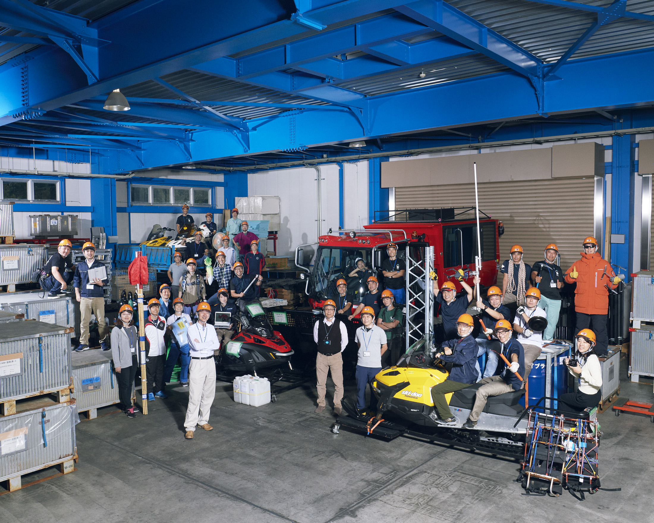 A large group of workers in hard hats pose together inside an industrial warehouse with vehicles and equipment around them.