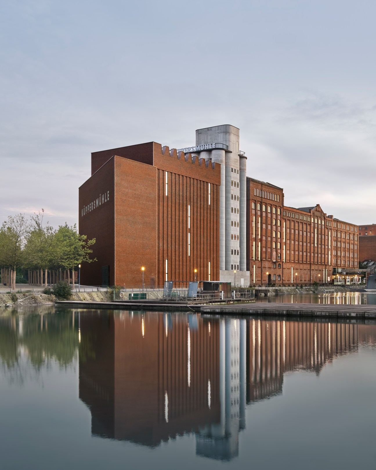 Modern brick museum extension with cylindrical concrete tower reflected in waterfront canal at dusk