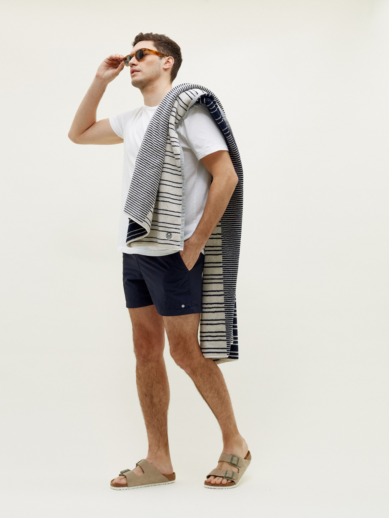 Man wearing white t-shirt and navy shorts holding striped beach towel and sunglasses