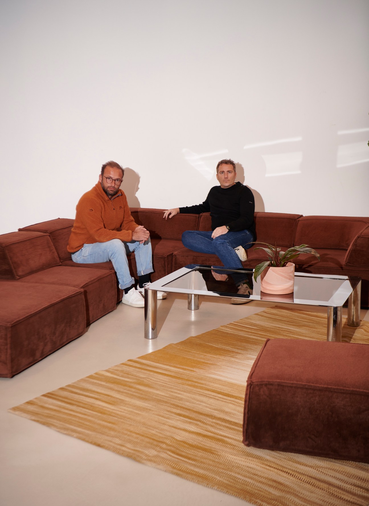 Two men sitting on a brown sectional sofa in a modern office space with white walls and a glass coffee table