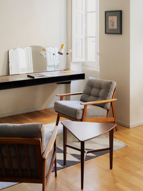 Modern hotel room with mid-century furniture, vanity desk with mirror and yellow lamp, wooden armchair and side table.