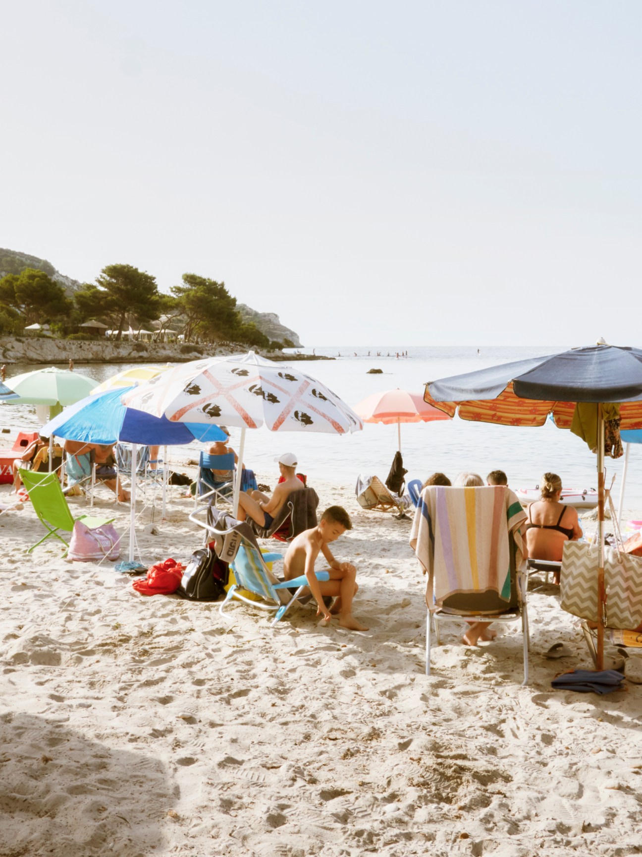 Beachgoers relax under colorful umbrellas on Calamosca beach with calm blue water and green hills in background