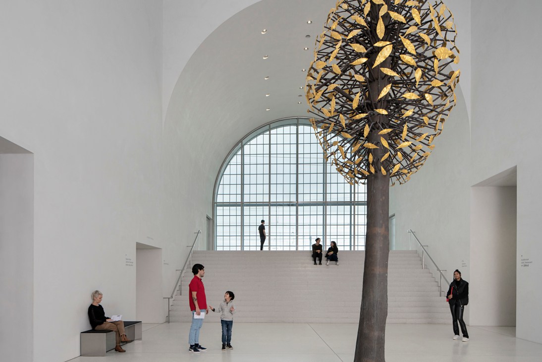 Large sculptural installation hanging from ceiling in white museum space with visitors viewing from stairs