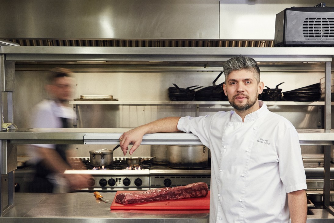 Polish chef Damian Wawrzyniak in white chef's coat standing in professional kitchen with raw meat on red cutting board
