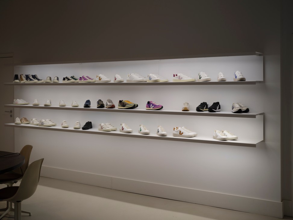 Display wall with three illuminated shelves showcasing various Veja sneakers in white, black and colorful designs